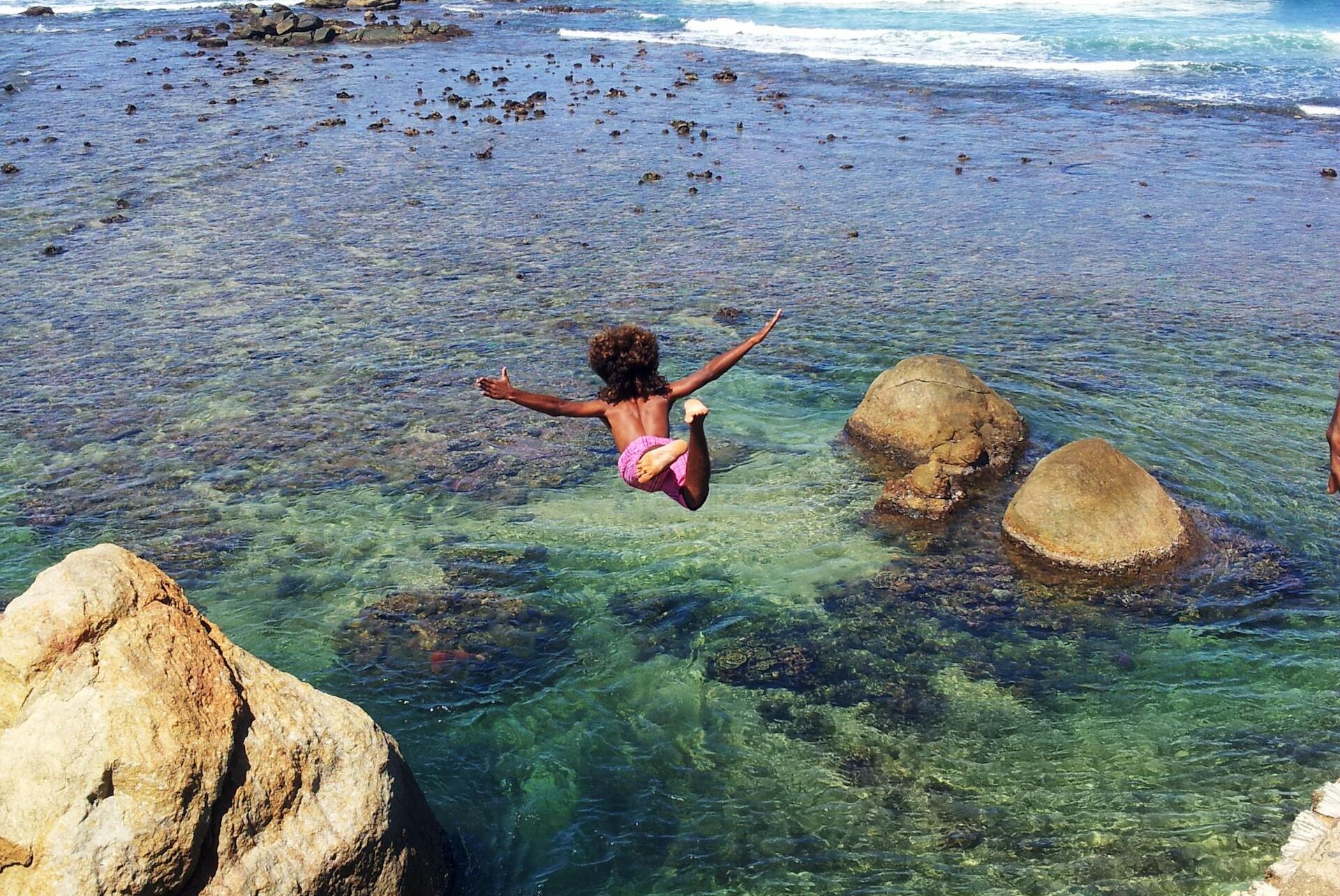 Person joyfully jumping into the clear ocean waters by the rocky Sri Lankan shore, embracing summer thrills.